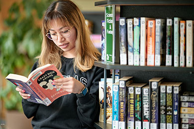 Student standing by leisure reading shelves and reading a book